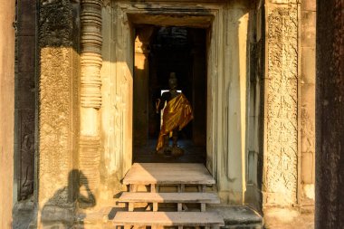 Ilık güneş ışığı Angkor Vats giriş köşkünün karmaşık bir şekilde oyulmuş kumtaşı kapı girişini aydınlatarak altından bir cübbeye sarılı yıpranmış Buda heykelini ortaya çıkarır. Tarihi Khmer mimarisi, dokulu kabartmalar ve derin gölgelerle vurgulanır.