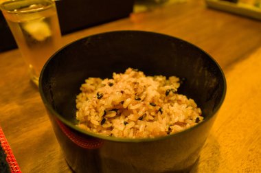 Japon çoklu tahıllı pirinci, siyah susam tohumlarıyla sunulur. Ahşap bir masada, sıcak ortam aydınlatmasıyla aydınlanan siyah bir kabın içinde. Sıcak bir yemek havası için..