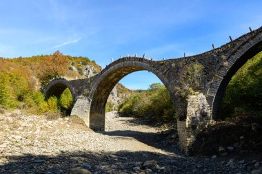 Tarihsel üçlü kemerli Plakidas Köprüsü, Yunanistan 'ın Epirus kentindeki Kipoi yakınlarında sonbahar yaprakları ve engebeli tepelerle çevrili kuru, kayalık bir nehir yatağını kaplıyor. Açık gün ışığı desenli taş işçiliğini ve Pindus Dağları 'nın canlı manzarasını gözler önüne seriyor..