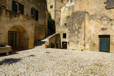 Matera, İtalya 'daki güneş taşı meydanı, yeşil kepenkleri ve saksı bitkileri olan yıpranmış tarihi binalarla çevrili. Kireçtaşı yüzeyler ve yumuşak gölgeler huzurlu bir Akdeniz atmosferini çağrıştırıyor..