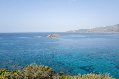 Capo Carbonara açıklarında küçük kayalık bir adanın etrafını çevreleyen canlı turkuaz sular, uzak dağlar ve parlak gökyüzü huzurlu bir Akdeniz manzarası yaratıyor..