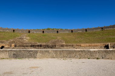 İtalya 'nın Pompeii kentindeki tarihi Roma amfitiyatrosunda katmanlı taş koltuklar ve çimenli setler yükseliyor. Açık mavi gökyüzü ve dokulu volkanik duvarlar, anıtların açık hava yapısını ve kalıcı mimari tasarımı ön plana çıkarıyor..