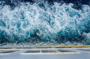 Akdeniz 'de çalkalanan canlı turkuaz ve beyaz köpüklü gemi güvertesi perspektifi. Kıtır su dokuları ve geometrik güverte çizgileri çarpıcı bir kontrast oluşturur.