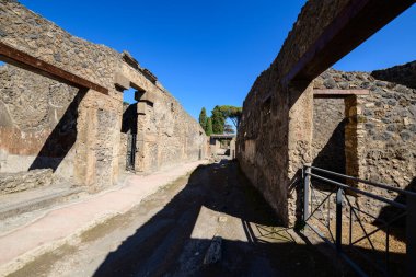 Pompeii, İtalya 'nın arkeolojik bölgesindeki yıpranmış volkanik taş ve tuğla duvarlarla çevrili dar Roma geçitleri. Parlak güneş ışığı keskin gölgeler bırakır, pürüzlü duvar dokularını ortaya çıkarır ve tarihi kalıntıların üzerinde parlak mavi bir gökyüzü.