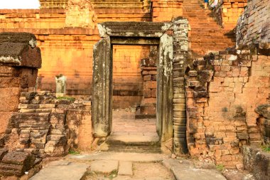 Kamboçya 'daki Pre Rup Tapınağı' nın dokulu ve kırmızı tuğlalı duvarları tarafından çerçevelenmiş yıpranmış taş bir kapı. Sıcak akşam güneşiyle aydınlatılmış. Sahne, aşınmış yüzeyler, tarihi mimari ayrıntılar ve yükselen tapınağın arka planının altını çiziyor.