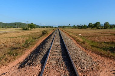 Kamboçya, Kamboçya 'da uzun ve düz demiryolu hatları kurak alanlardan ve kırsal alanlardan geçer. Sahnede açık mavi gökyüzü, dağınık ağaçlar, uzak yeşil tepeler ve parlak gün ışığı altında elektrik direkleri yer alıyor..