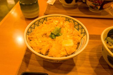 Taze yeşil garnitürle kaplanmış bir kase Japon tavuğu ve yumurtalı donburi sıcak ortam aydınlatmasında ahşap bir tepside oturur. Parlak yumurta ve taze tavuk rahatlatıcı, davetkar bir sunum oluşturur..