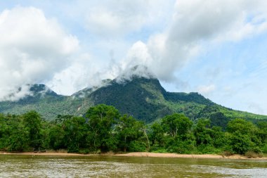 Kuzey Laos 'taki Nam Ou Nehri üzerindeki yemyeşil bir dağın zirvelerinde beyaz bulutlar yüzer. Yoğun tropikal orman, parlak bir gökyüzünün altında canlı bir manzara oluşturan nehir kıyısı boyunca uzanır..