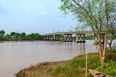 Renkli bayraklı beton bir köprü Ben Tre, Mekong Delta, Güney Vietnam 'da geniş ve çamurlu bir nehir boyunca uzanır. Sahnede nehir kıyısı boyunca yeşil ağaçlar, elektrik direkleri ve yumuşak gün ışığı altında çimenli bitki örtüsünün arasında demirlenmiş ahşap bir tekne yer alıyor..