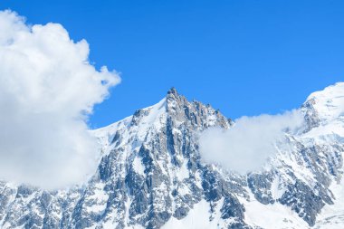 Aiguille du Midi 'nin karla kaplı zirvesi Fransız Alpleri' ndeki yumuşak beyaz bulutlarla kısmen örtülü açık mavi gökyüzüne doğru hızla yükselir..
