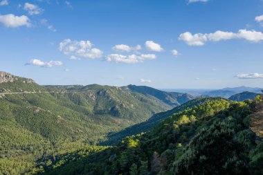 Sardunya 'daki Baunei ve Dorgali yakınlarındaki gür orman dağlarının geniş bir manzarası, dağ sırtları boyunca görünen dolambaçlı bir yol ve parlak gökyüzünü noktalayan dağınık bulutlar..