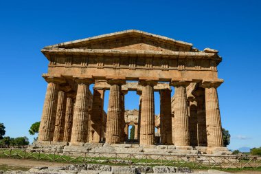 İtalya 'nın Paestum kentindeki Neptün Tapınağı' nın doğrudan bir perspektifi, açık mavi gökyüzünün altında devasa Doric sütunlarını ve yıpranmış taş cephelerini sergiliyor. Antik Yunan yapısı, kırsal ahşap çitlerle çevrili, yükseltilmiş bir platformda duruyor.