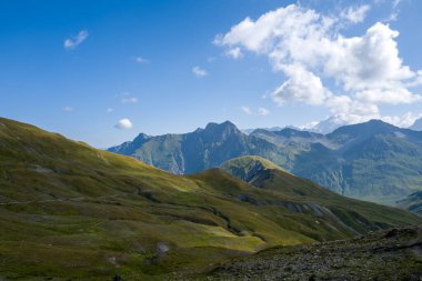 Güneşli çimenli yamaçların geniş manzarası engebeli dağlara doğru uzanır parlak mavi bir gökyüzünün altında Val dAoste bölgesinde dağınık bulutlar.