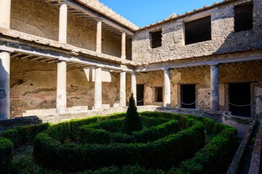 Manikürlü boxwood çitleri ve merkezi bir çalı olan güneşli bir avlu, Pompeii, İtalya 'daki Aşıklar Evi' ndeki antik taş sütunlar tarafından çerçevelenmiştir. Yıpranmış duvarlar ve solmuş fresk izleri geometrik bahçenin etrafını sararak sakin bir tarihi eser yaratıyor.