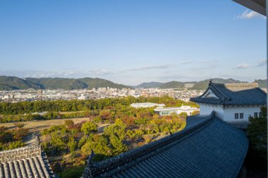 Himeji Şatosu 'nun süpürücü manzarası süslü kiremitli çatılar, sonbahar ağaçlarının renkli bir karışımı, ve şehir manzarası açık öğleden sonra ışığı altında yeşil dağlara karşı..
