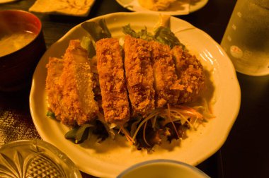 Japon Hongu 'da sıcak ortam aydınlatması altında seramik tabakta sunulan karışık salata yeşilliklerinden oluşan bir yatakta servis edilen altın ekmekli domuz pirzolası. Sahne çıtır çıtır dokuları ve davetkar yemek cazibesini ön plana çıkarıyor.