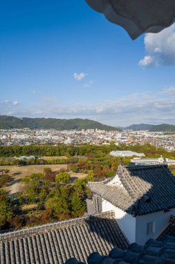 Himeji Kalesi 'nden yükselen manzarada ön planda geleneksel fayanslı çatılar, geniş şehir manzarası ve dağınık mavi bulutlu canlı bir gökyüzü altında yemyeşil parklar yer alıyor..