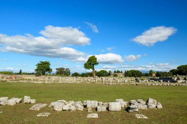 İtalya 'nın Paestum kentindeki arkeoloji sahasında dağılmış antik taş blokları ve yalnız ağaçlarla geniş bir çim forumu. Sahne, dramatik beyaz bulutlar ve ufukta görünen uzak tapınak sütunlarıyla canlı bir mavi gökyüzünün altında geçiyor..