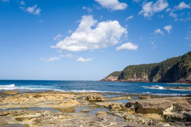 Katmanlı kaya oluşumları Goshikihama Sahili 'nin kıyıları boyunca uzanır. Derin mavi dalgalar ve dağılmış bulutlarla dolu parlak bir gökyüzünün altında orman burnu vardır..
