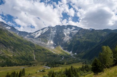 Muhteşem buzullar engebeli dağ yamaçlarından aşağı süzülüyor. Dağılmış ağaçlar ve sarmal dereleri olan yeşil bir vadiye bakıyor. Fransız Alpleri 'ndeki parçalı bulutlu bir yaz gökyüzünün altında..