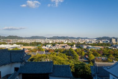 Himeji Kalesi 'nin geniş açılı görüntüsü kiremitli çatıları, yemyeşil parkları ve açık mavi gökyüzünün altındaki uzak dağlarla genişleyen Himeji şehrini gösteriyor..