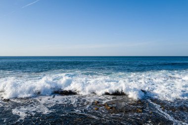 Kotohikihama Sahili 'nde kayalık bir kıyıya vuran parlak mavi dalgalar, açık bir gökyüzünün altında beyaz köpüğü aydınlatıyor..