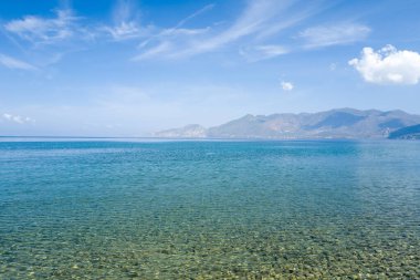 Yüzeyin altında pürüzsüz çakıl taşları olan şeffaf sığ deniz, Psifta sahilinde, Peloponnese, Argolide 'de, parlak bir gökyüzü altında, geniş ve sakin bir mavi suya ve engebeli dağlara açılıyor..