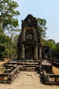 Camları açık ve karmaşık oymaları olan uzun kumtaşı kule, Kamboçya Angkor bölgesindeki Preah Khan tapınağının ana geçidinde yükseliyor. Tropikal ağaçlarla çevrili yıpranmış taş yapı, tarihi Khmer 'i harmanlayan açık gökyüzünün altında duruyor.