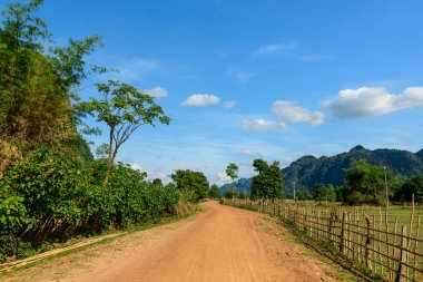 Kırmızımsı toprak yol, bambu çitleri ve yemyeşil çimenler arasında yavaşça kıvrılır ve dağınık bulutlu, parlak mavi bir gökyüzünün altındaki uzak kireçtaşı tepelerine çıkar. Sakin kırsal kesimde açık alanlar, kırsal çitler ve doğal çitler yer almaktadır.