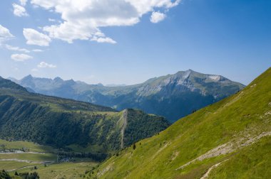 Bereketli yeşil tepeler ve engebeli tepeler berrak mavi bir gökyüzünün altında, yaz mevsiminde güneşli dağların üzerinde süzülen yumuşak bulutlarla süpürülüyor..