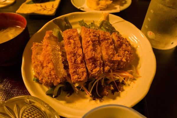 Japon Hongu 'da sıcak ortam ışıklarıyla aydınlatılan seramik bir tabakta karışık salata sebzeleri ve doğranmış sebzelerden oluşan bir yatakta dilimlenmiş ve dilimlenmiş gevrek domuz pirzolası. Sahnede çıtır çıtır dokular ve davetkar sunumlar vurgulanıyor.