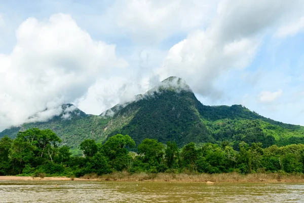 Kuzey Laos 'taki Muang Ngoi yakınlarındaki Nam Ou Nehri' nin üzerinde yükselen yoğun ormanlık bir dağda beyaz sis sürükleniyor. Yeşiller yemyeşil ağaçlar ve yoğun bitki örtüsü... dramatik bulutlarla dolu bir gökyüzünün altındaki nehir kıyısı..