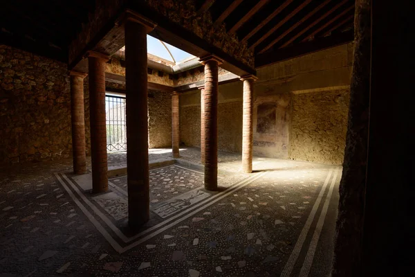 Tuğla sütunları ve açık tavan pencereli bir Roma avlusu İtalya 'nın Pompeii kentindeki Menander Evi' nin geometrik mozaik zeminini aydınlatıyor. Güneş ışığı, bu arkeolojik alanın dokulu taş duvarlarını ve tarihi mimari ayrıntılarını vurguluyor.