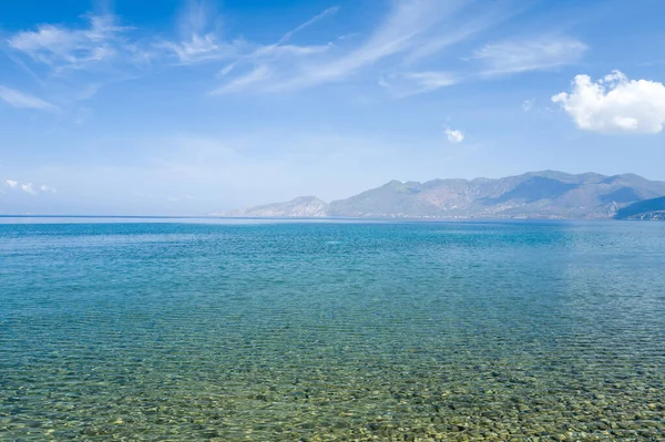 Yüzeyin altında pürüzsüz çakıl taşları olan şeffaf sığ deniz, Psifta sahilinde, Peloponnese, Argolide 'de, parlak bir gökyüzü altında, geniş ve sakin bir mavi suya ve engebeli dağlara açılıyor..