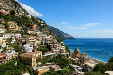 Rengarenk yamaçlı villalar ve sembolik sarı kubbeli kilise, İtalya 'nın Positano kentindeki Amalfi Sahili' nin turkuaz sularına bakıyor. Güneşli cepheler, yemyeşil bahçeler ve dramatik uçurumlar canlı bir Akdeniz köyü manzarası yaratıyor.