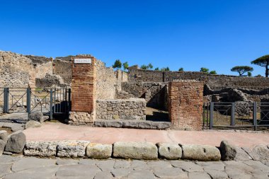 Yıpranmış tuğla sütunlar ve volkanik taş duvarlar tarafından çerçevelenmiş tarihi bir giriş, İtalya 'nın Pompeii arkeoloji sahasında açık mavi bir gökyüzünün altında duruyor. Sahne pürüzlü duvarcılık, demir korkuluklar ve antik çağların ayırt edici dokularını vurgular.