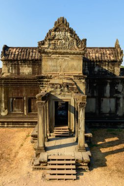 Kamboçya 'nın Angkor Vat kentinin ikinci katındaki zengin kumtaşı giriş pavyonunun detaylı görüntüsü. Sıcak güneş ışığı, kuru çimlere ve açık mavi gökyüzüne karşı, karmaşık çatı çizgisi, yıpranmış sütunlar ve desenli taş işçiliğini vurgular