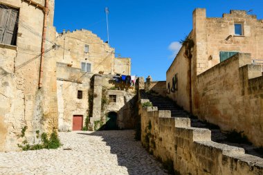 Matera, İtalya 'daki antik taş geçitte yıpranmış kireçtaşı binalar, kırsal bir merdiven ve canlı mavi bir gökyüzü altında dokulu cepheler bulunuyor. Güneş ışığı, tarihi Akdeniz karakterinin altını çizerek güçlü gölgeler bırakıyor.