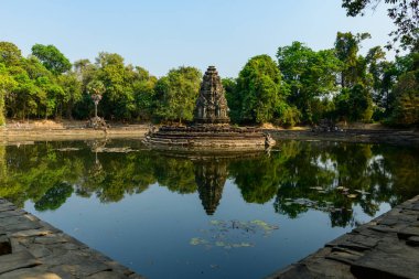 Neak Pean tapınağındaki sakin bir göletin merkezinden yükselen taş tapınak, verimli tropikal ağaçlar ve Kamboçya Angkor bölgesindeki sakin sularla çevrilidir. Tarihi duvarcılık ve berrak yansıma mavi gökyüzünün altında huzurlu bir atmosfer yaratır..
