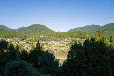 Yoğun yeşil ormanlar ve açık mavi gökyüzünün altındaki yuvarlanan tepelerle çevrili Chikatsuyu köyünün geniş bir manzarası var. Parlak güneş ışığı canlı manzarayı ve huzurlu kırsal manzarayı vurgular.