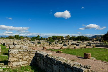 İtalya 'nın Paestum kentindeki arkeolojik alanda tarihi taş duvarlar ve tuğla sütunlar çimenli açık alanlar boyunca uzanıyor. Sahne, dağınık bulutlar ve uzak ağaçlarla dolu canlı, mavi bir Akdeniz gökyüzünün altında çekilmiştir.
