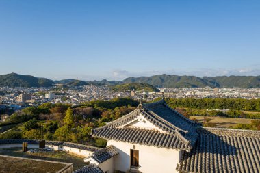 Himeji Kalesi 'nden yükseltilmiş perspektif ön planda geleneksel fayanslı bir çatı, canlı yeşil ağaçlar ve açık gün ışığı altında yuvarlanan dağlara karşı yayılan şehir manzarası..
