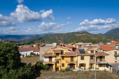 Terracotta çatıları olan parlak binalar Orgosolo 'nun kıyısında duruyor, yuvarlanan yeşil tepelere ve dağınık mavi bulutların altında uzak dağlara bakıyor..