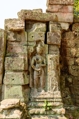 Kamboçya 'nın Angkor bölgesindeki Preah Khan tapınağındaki yıpranmış, liken kaplı bir duvara, ayakta duran bir tanrının ayrıntılı kumtaşı heykeli yerleştirildi. Karmaşık rahatlama ve yaşlanmış taş bloklar, tapınağı aydınlatan sıcak gün ışığıyla aydınlatılıyor.