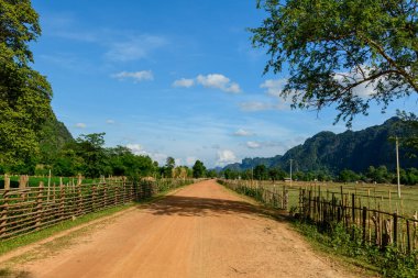 Kırsal bambu çitleriyle kaplı düz toprak bir yol açık araziler ve yeşillikler arasında uzanır, canlı mavi gökyüzünün altındaki kireçtaşı tepeleri tarafından çerçevelenir. Sahne, Laos kırsalının sakin ve güneşli atmosferini doğal bir şekilde yakalıyor.