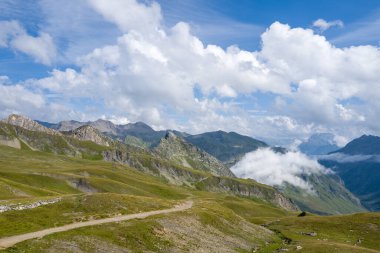 Dolambaçlı bir toprak yol otlak bir dağ sırtından geçer dramatik dağ zirveleri ve yuvarlanan bulutlar parlak mavi bir gökyüzü altında özgürlük ve enginlik hissi uyandırır..