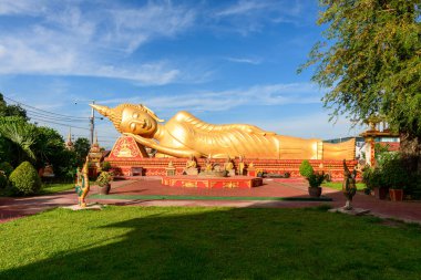Luang in Vientiane, Laos 'taki Pha' da uzanan altın sarısı Buda heykeli parlak sabah güneşiyle aydınlanır. Manikürlü çimenler, süslü kırmızı kaideler ve tropikal yeşilliklerle çevrili sakin figür canlı mavi gökyüzüne karşı duruyor.