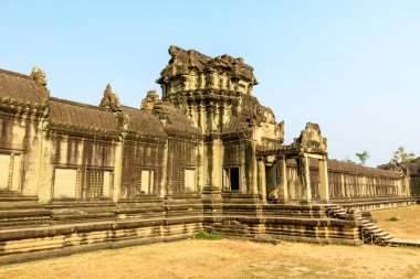 Sıcak öğleden sonra güneşi, yıpranmış kumtaşı galerisini ve Angkor Vat, Kamboçya 'nın merkez kulesini yıkıyor. Karmaşık Khmer mimarisi, tarihi ihtişam ve tropikal manzarayı vurgulayan kuru çimenler ve açık mavi bir gökyüzü tarafından çerçevelenmiştir..