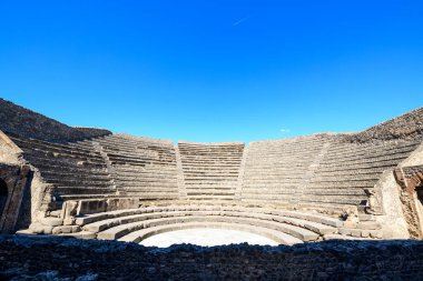 Yarı dairesel yıpranmış taş koltuklar İtalya 'nın Pompeii kentindeki tarihi küçük tiyatroyu oluşturur. Parlak güneş ışığı, amfitiyatronun geometrik çizgilerini canlı, bulutsuz gökyüzüne vurgular..