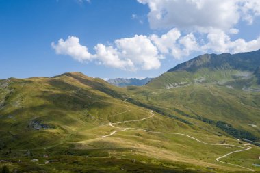 Dolaşan dağ yolları, Col de Balme bölgesinde dağınık bulutlarla birlikte parlak mavi bir gökyüzünün altında yuvarlanan çimenli yamaçlar boyunca esiyor. Güneş ışığı ve gölge açık arazide dinamik dokular yaratır..